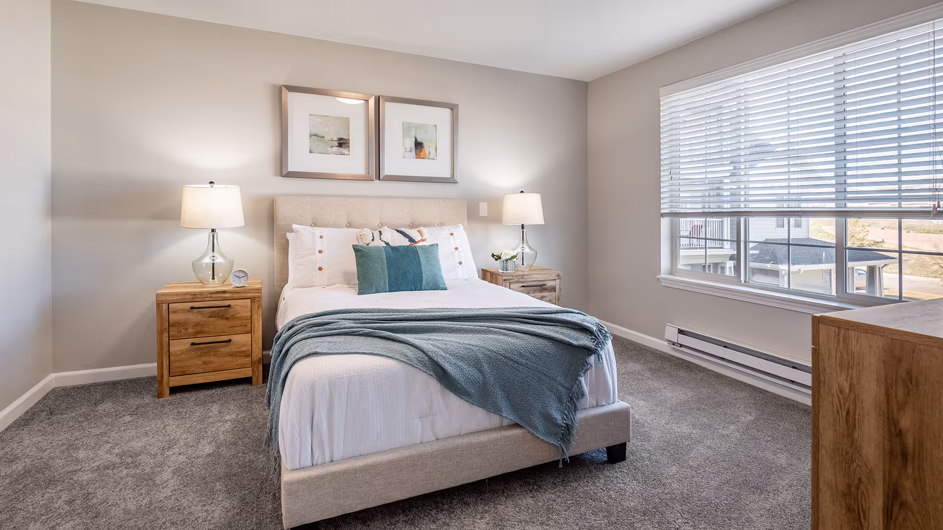 A bright and cozy bedroom with a neatly made bed featuring white linens, a teal throw blanket, and decorative pillows. There are two wooden nightstands on either side of the bed, each with a glass lamp and small decor items. Above the bed are two framed abstract art pieces. A large window with white blinds allows natural light to fill the room, and the floor is carpeted in gray.