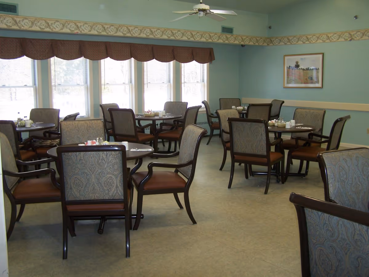 A dining room with several round tables and cushioned chairs arranged neatly. The room has light blue walls, a decorative border near the ceiling, and a ceiling fan. Large windows with valances allow natural light to fill the space. A framed picture hangs on one wall.