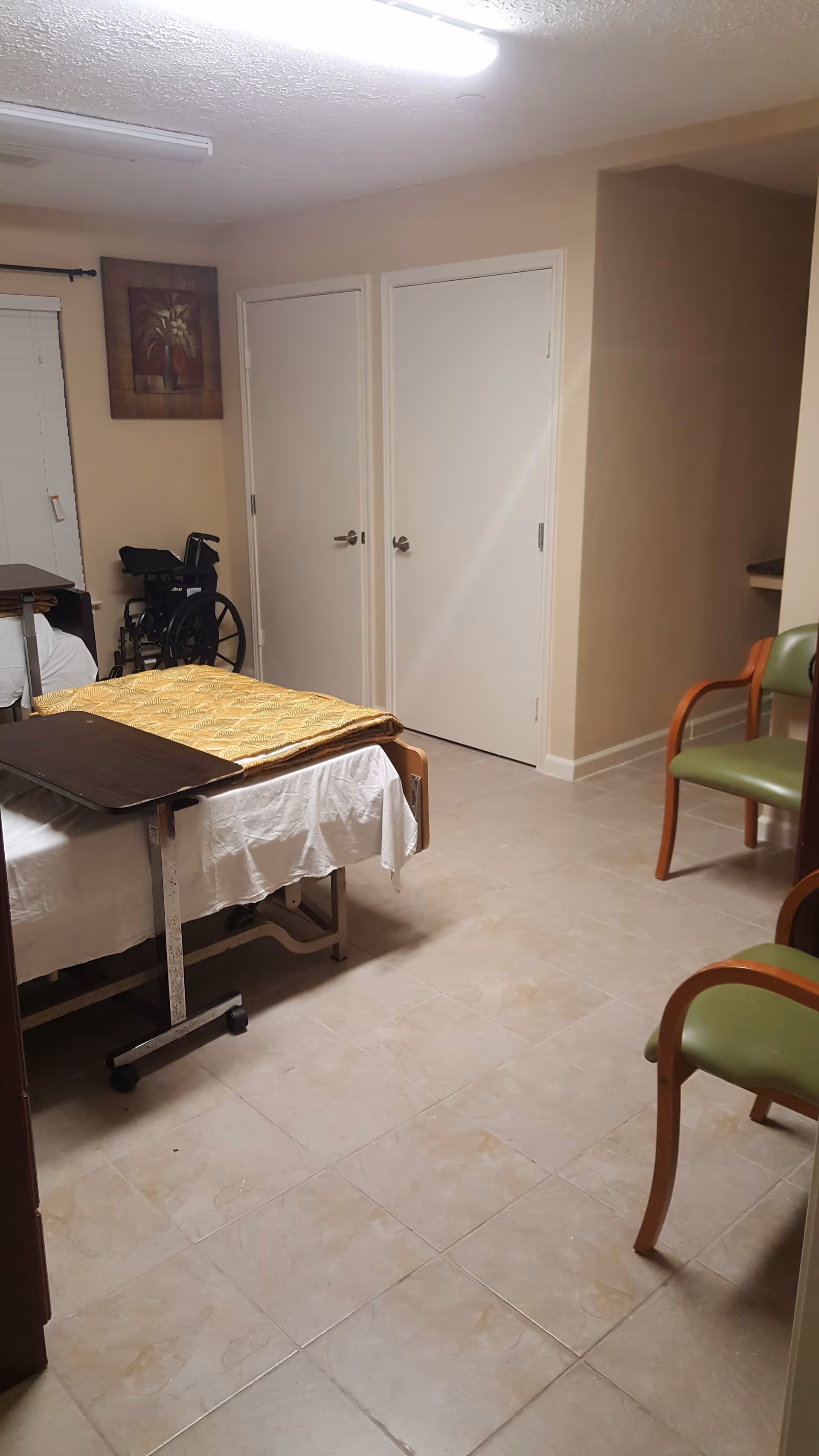 Assisted-living resident room with a hospital bed and overbed table, a wheelchair, two closed doors, and seating chairs.