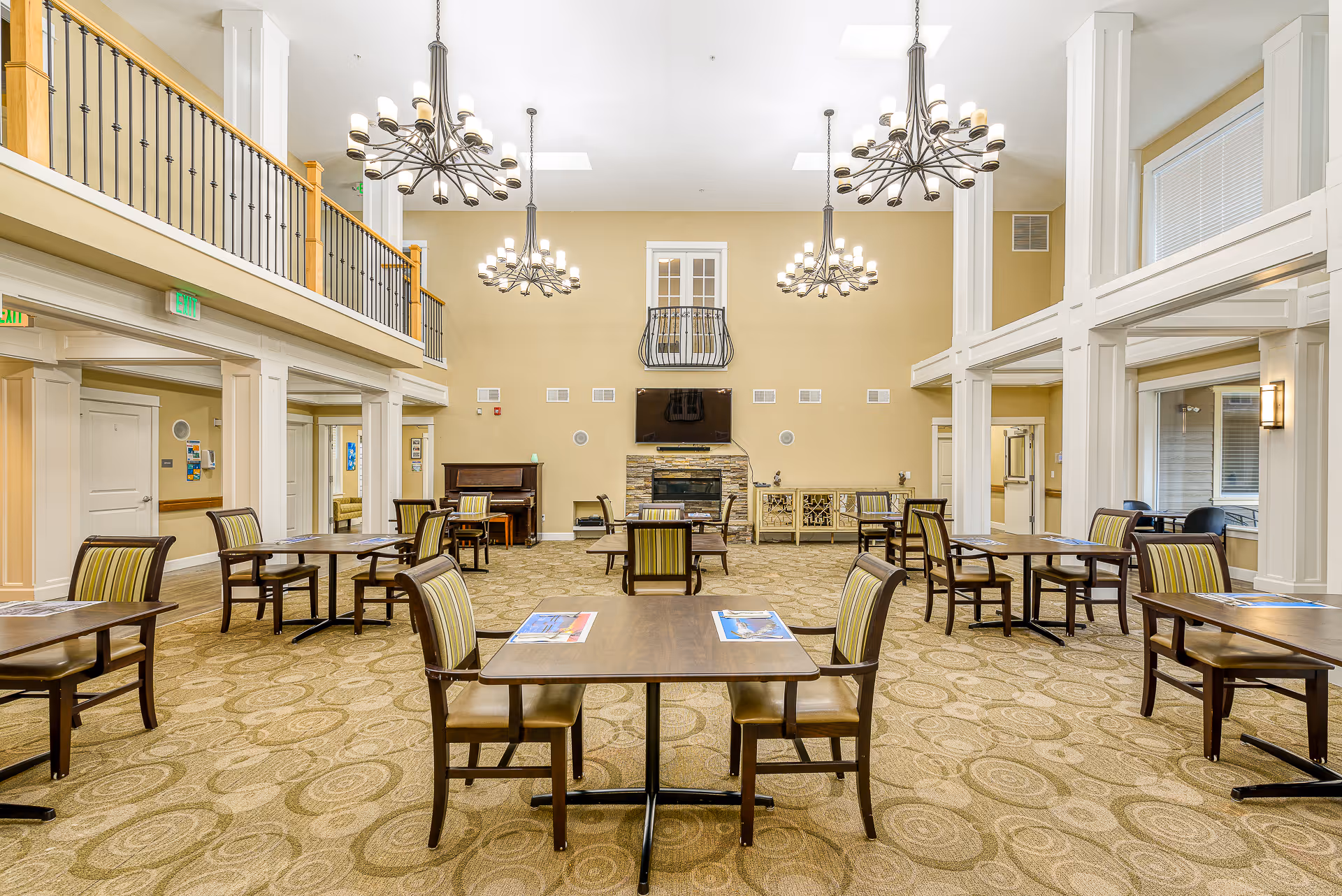 Spacious and well-lit dining room in a senior living facility with multiple wooden tables and chairs arranged neatly on a patterned carpet. The room features high ceilings with three large chandeliers, a fireplace with a mounted TV above it, and a piano in the corner. There is a balcony railing on the upper level and large windows allowing natural light to enter.