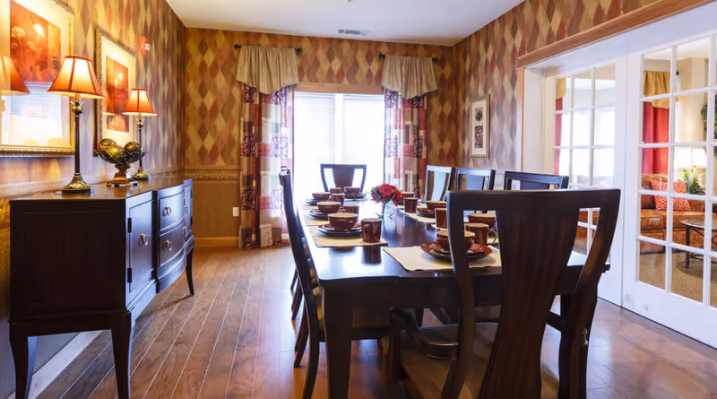 Dining room with a dark wood table and chairs set for a meal, a matching sideboard, patterned wallpaper, and French doors to a sitting area.