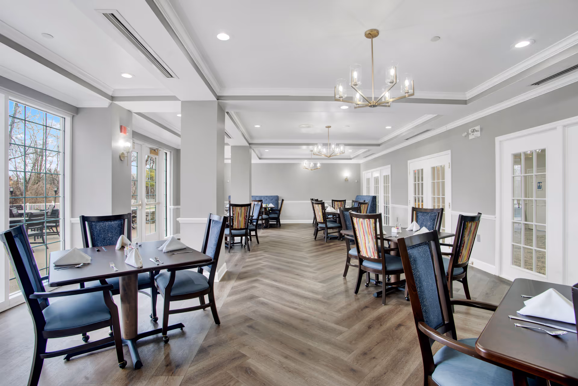 A bright and spacious dining room with multiple dark wood tables and chairs, each table set with white folded napkins and silverware. The room features large windows and glass doors letting in natural light, light gray walls, wood flooring in a herringbone pattern, and modern chandeliers hanging from the ceiling. Outside the windows, there is a patio area with additional seating.