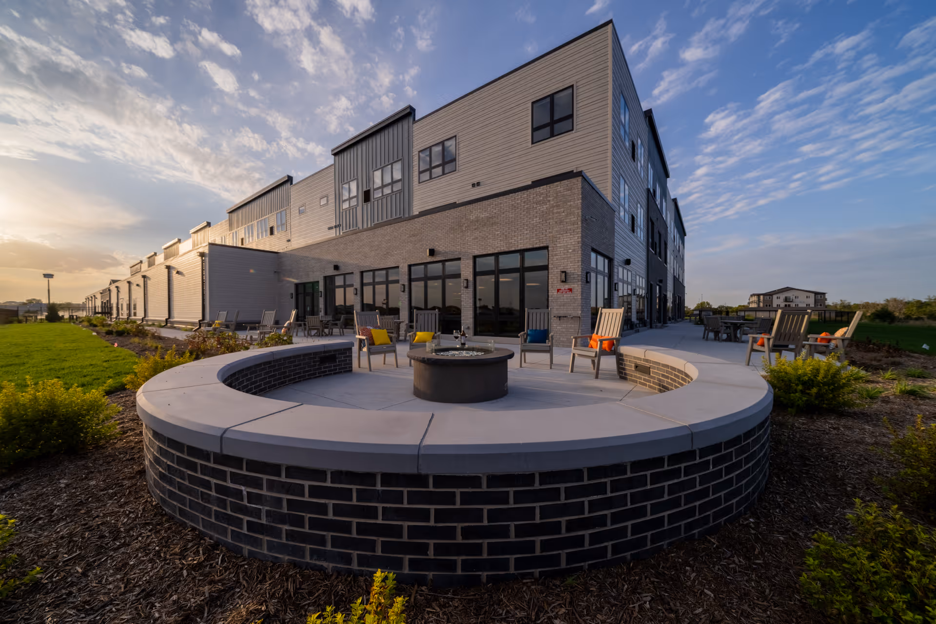 Outdoor patio area at Salt Creek Senior Living featuring a circular brick fire pit surrounded by chairs with colorful cushions, adjacent to a modern multi-story building under a partly cloudy sky during sunset.