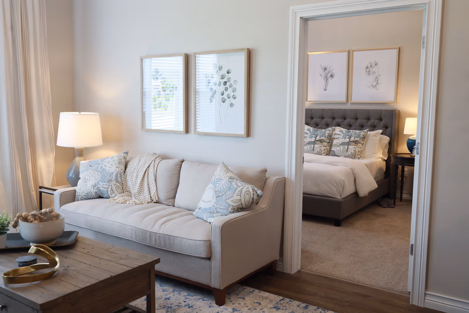 Bright living room with a beige sofa, patterned pillows, a wooden coffee table and a lamp, looking into a bedroom with a tufted bed.