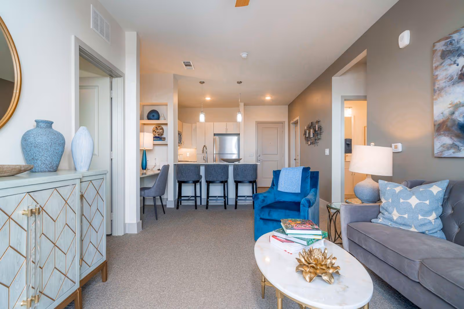 A modern living room and kitchen area in a senior living facility. The living room features a gray sofa with a blue patterned pillow, a blue armchair with a light blue throw, and a white oval coffee table with books and a decorative golden flower. The kitchen has white cabinets, a stainless steel refrigerator, and a counter with three gray bar stools. There is a small desk area with a gray chair and a lamp, and the walls are painted in neutral tones with some artwork and decorative items.