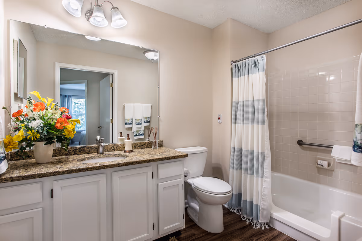A clean and well-lit bathroom featuring a white toilet, a bathtub with a striped shower curtain, a granite countertop with a sink, and a large mirror above the sink. There is a vase with colorful flowers on the countertop and neatly folded towels hanging on the wall. The floor has a wood-like finish.