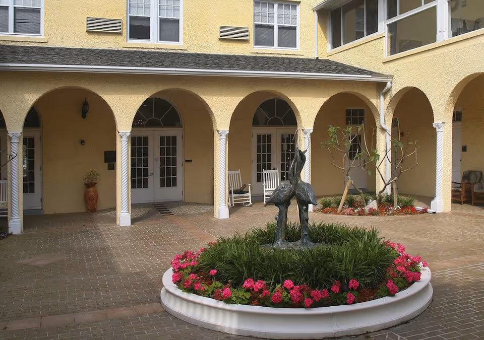 Courtyard area of a senior living facility with a circular flower bed featuring a sculpture of two birds, surrounded by pink flowers and greenery. The courtyard is paved with bricks and has a yellow building with arched doorways and white columns in the background. There are white rocking chairs and potted plants near the doors.