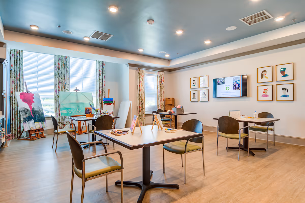 Bright community art and activity room with tables, chairs, easels and paintings near large windows.
