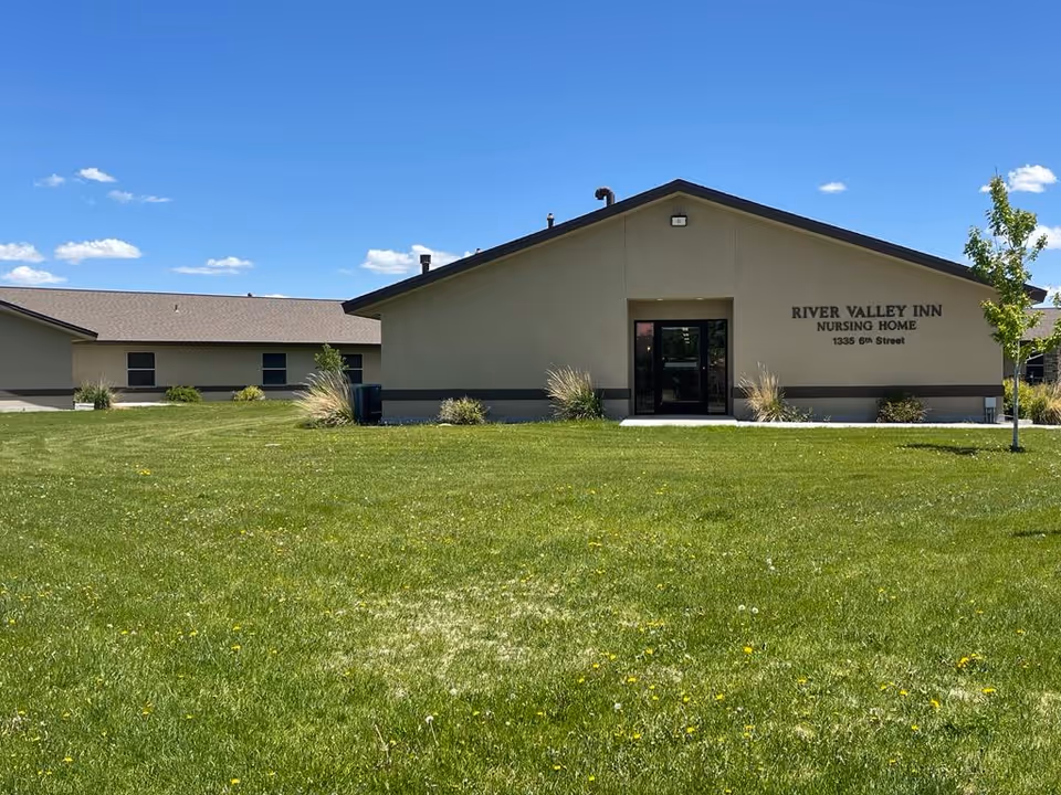 Exterior view of a single-story beige building with a dark roof under a clear blue sky. The building has a sign that reads 'RIVER VALLEY INN NURSING HOME 1335 6th Street'. There is a large grassy lawn in front with a few small plants and a young tree on the right side.