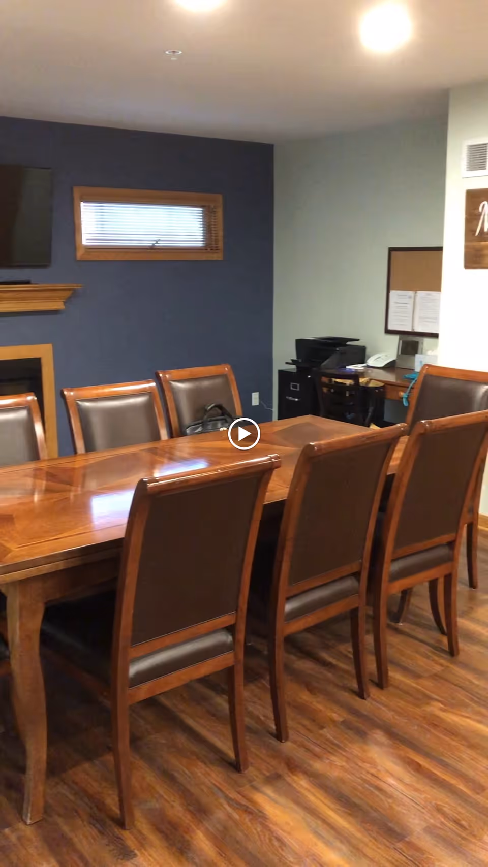 A dining room with a long wooden table surrounded by eight wooden chairs with brown cushioned seats and backs. The room has wood flooring, a dark blue accent wall with a small window and a mounted TV above a fireplace. In the corner, there is a small desk area with office equipment including a printer and a corkboard on the wall.
