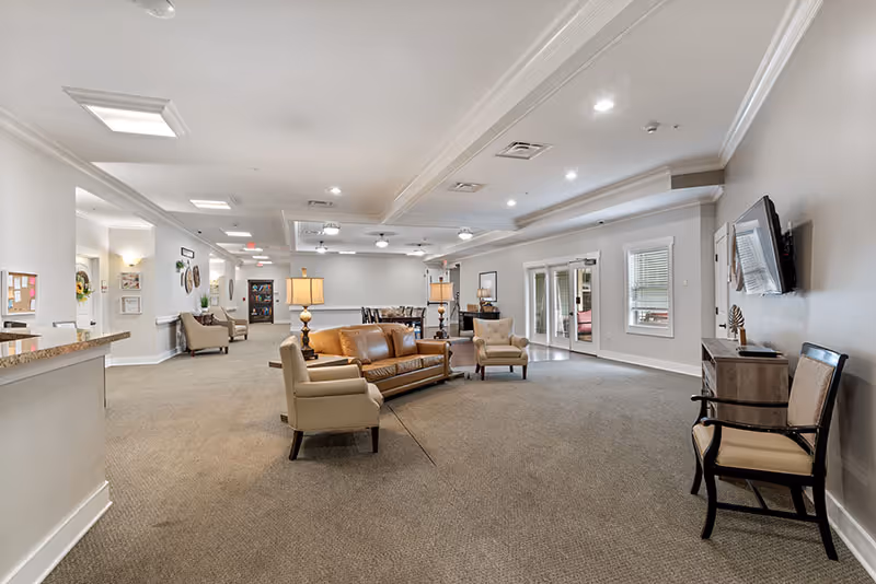 Spacious senior living facility common area with beige carpet, neutral walls, and recessed lighting. The room features a brown leather sofa, beige armchairs, two table lamps, a wall-mounted TV, and a small cabinet. In the background, there is a dining area with tables and chairs, and glass doors leading to another room.