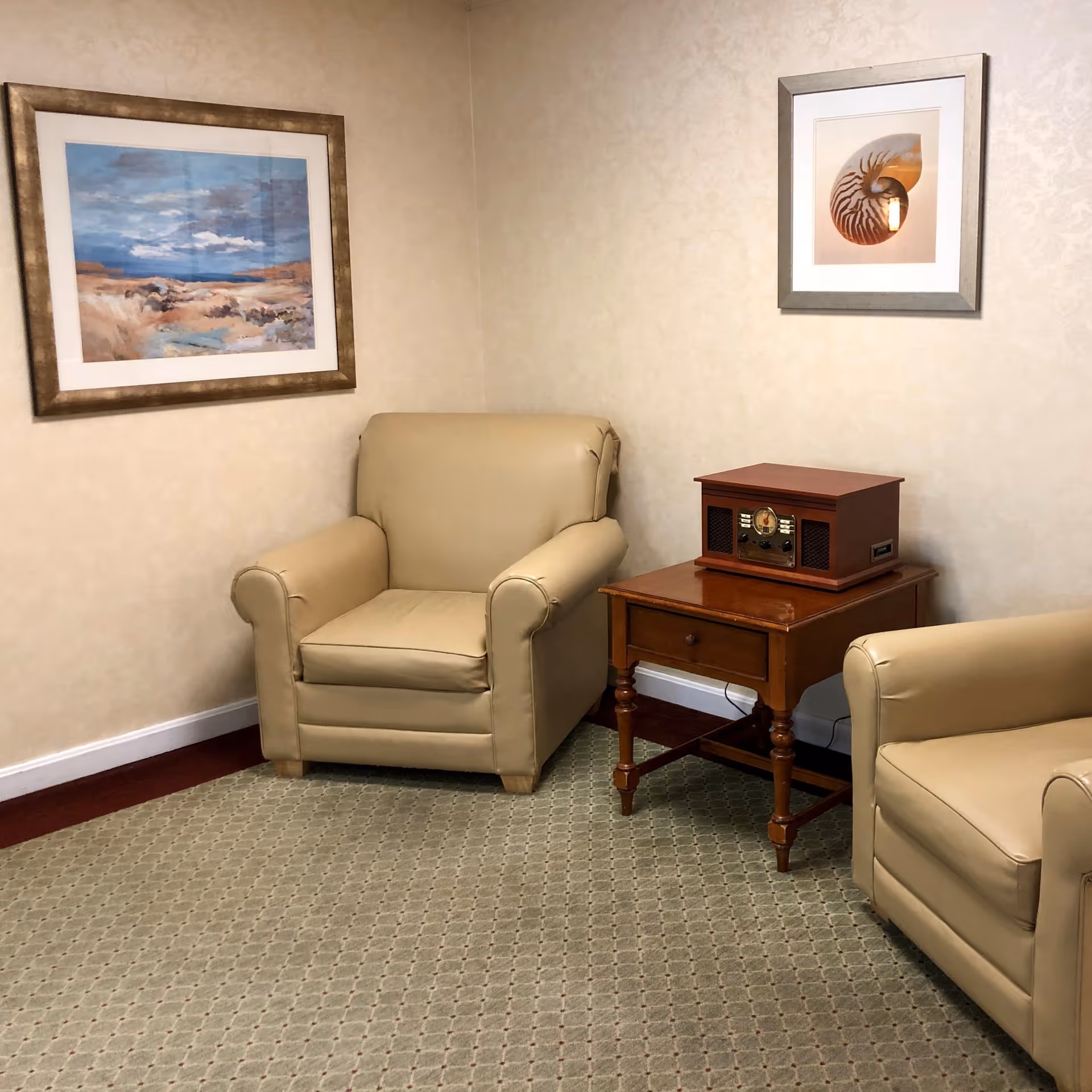 A cozy corner of a room with two beige armchairs placed on a patterned carpet. Between the chairs is a wooden side table with a vintage-style radio on top. The walls are light-colored with subtle patterns, and two framed pictures hang above the chairs, one depicting a landscape and the other a seashell.