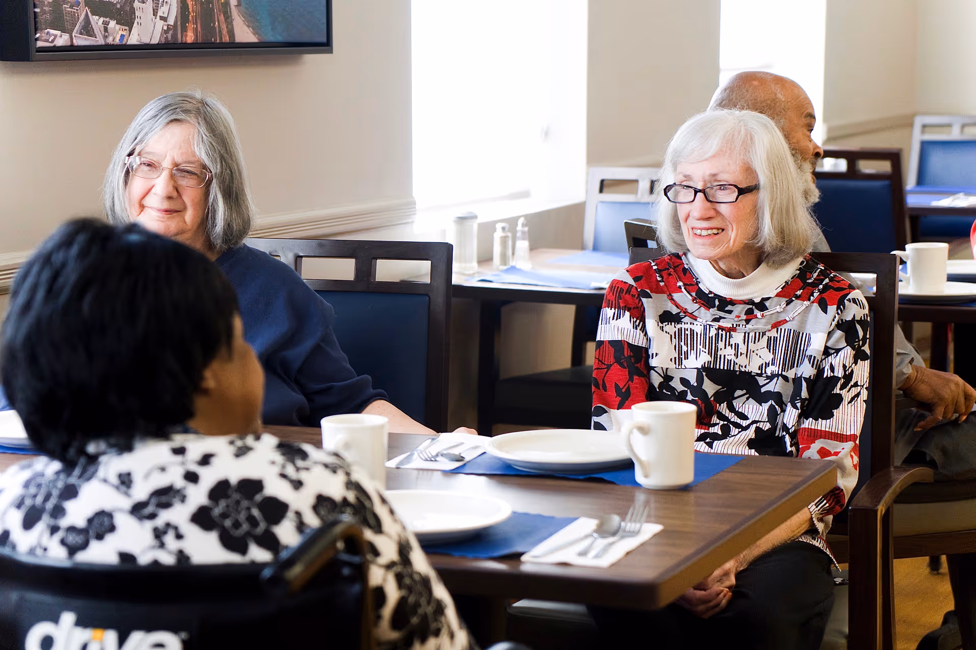 Several elderly residents sit and chat at a table in a communal dining room with plates and mugs set.