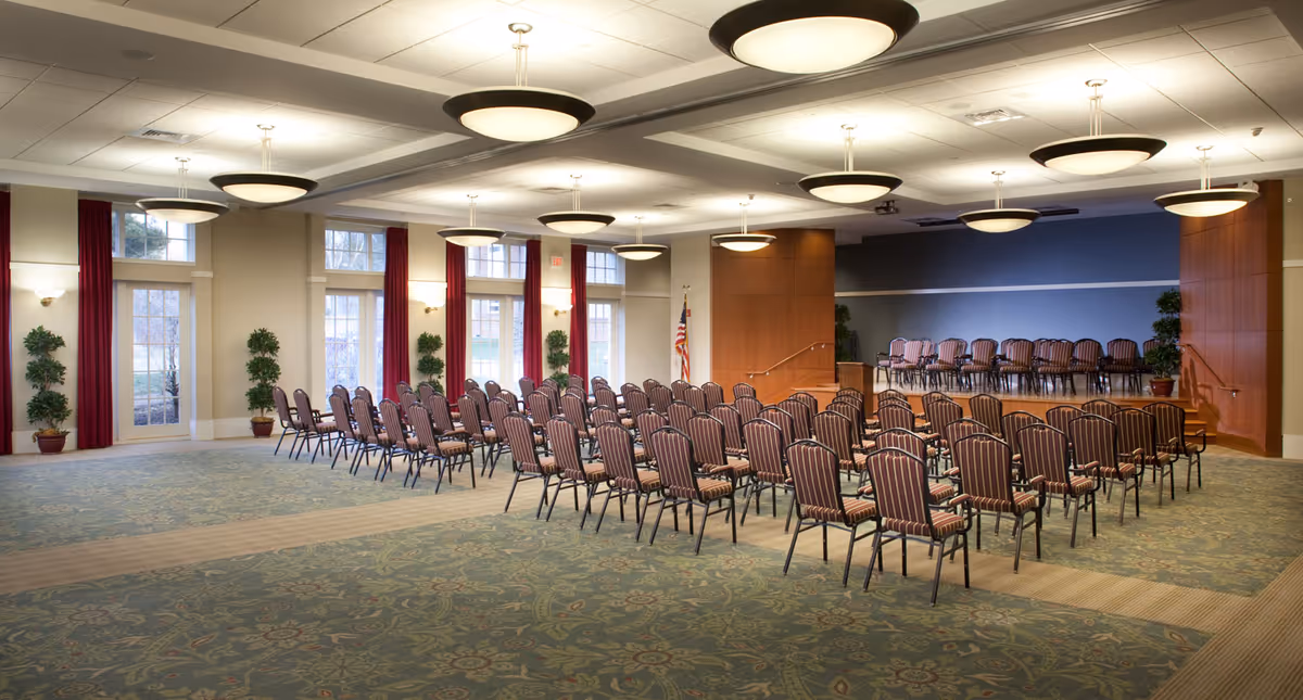 A large, well-lit meeting or event room with rows of striped chairs arranged facing a small stage area. The room has large windows with red curtains, several potted plants, and ceiling lights evenly spaced across the ceiling. An American flag is positioned near the stage.