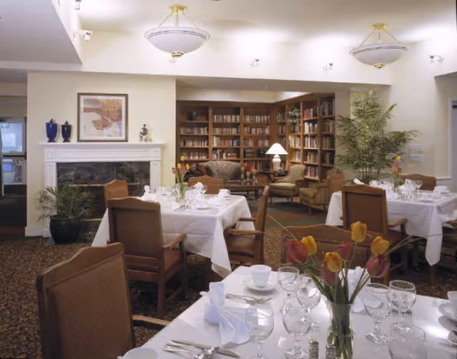 A cozy dining room with tables set for a meal, featuring white tablecloths, glassware, and floral centerpieces. In the background, there is a fireplace with decorative items on the mantle, a large bookshelf filled with books, comfortable armchairs, and a floor lamp. The room has warm lighting and a carpeted floor.