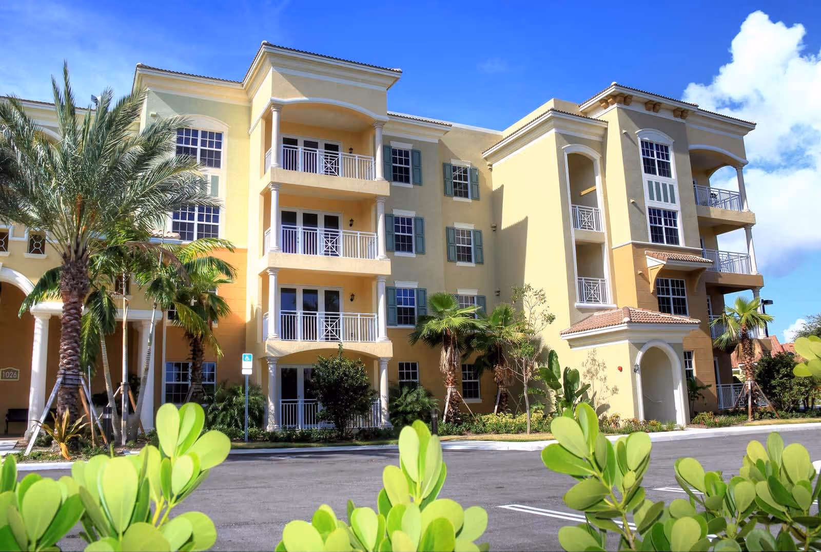 Exterior view of a multi-story senior living facility building with balconies, palm trees, and green shrubs under a blue sky with some clouds.