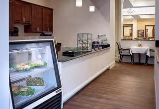 Interior view of a dining area in a senior living facility featuring a refrigerated display case with sandwiches and salads, a counter with food service items, and a small dining table with chairs in the background.