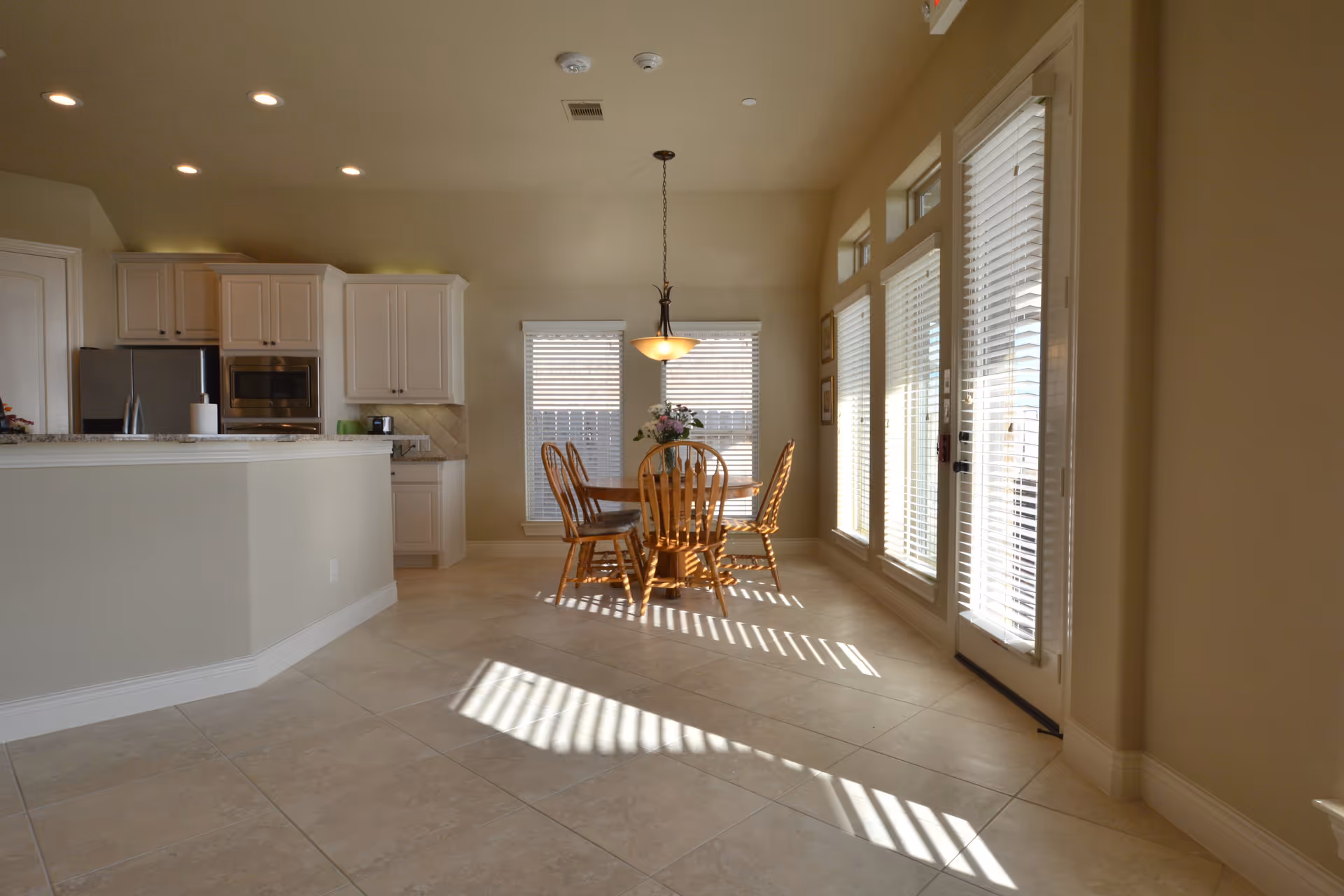 Bright dining area with a round wooden table and four matching chairs, a hanging light fixture above the table, large windows with white blinds letting in sunlight, and a kitchen area with white cabinets and stainless steel appliances in the background.