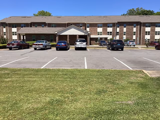 Front view of a two-story brick building with a covered entrance and several parked cars in front. The building is surrounded by a green lawn under a clear blue sky.