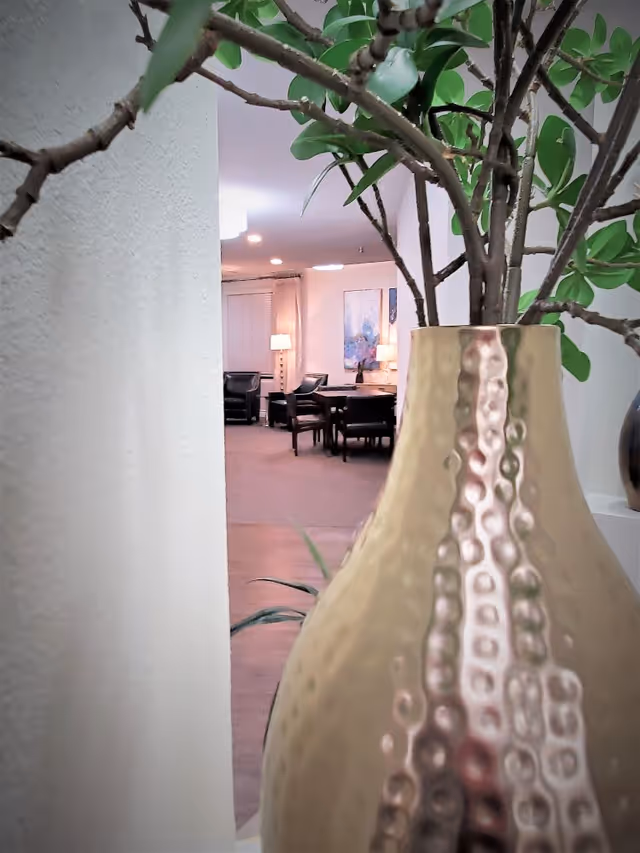 Close-up view of a large textured gold vase with green leafy branches, with a softly lit senior living facility common area in the background featuring chairs, tables, lamps, and artwork on the wall.