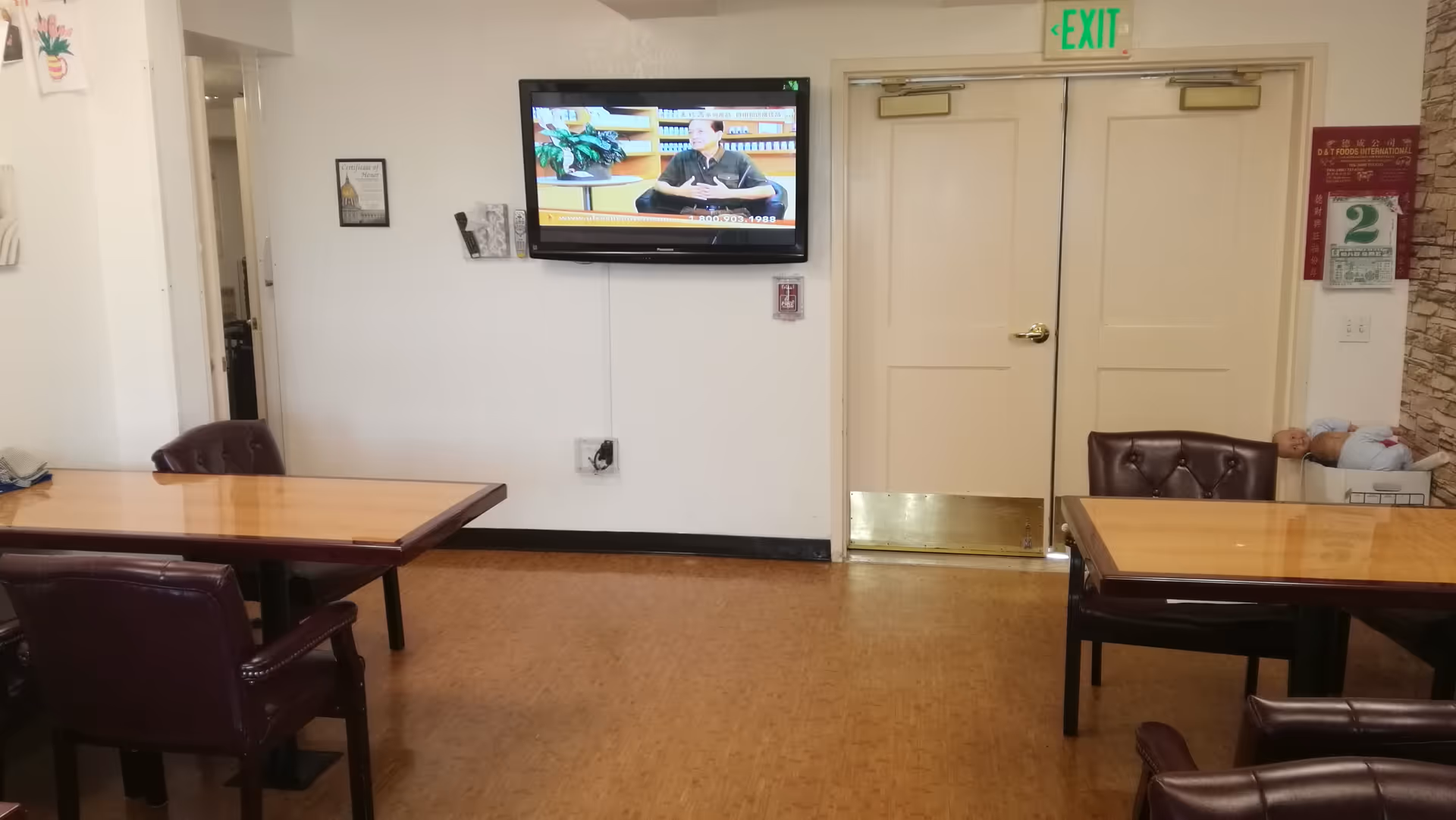 Interior room with wooden tables and dark leather chairs, a wall-mounted television displaying a man speaking, double doors with an exit sign above, and a box with two baby dolls in the corner.