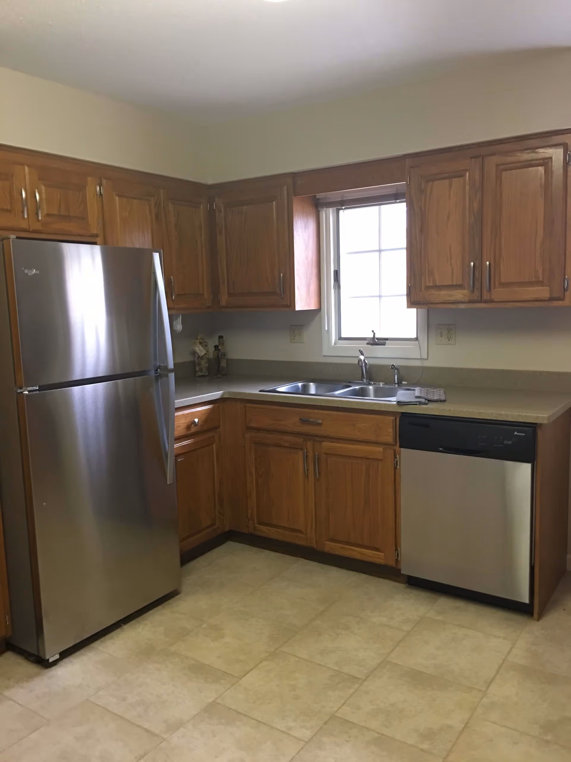 Small kitchen with stainless steel refrigerator and dishwasher, wooden cabinets, a double sink under a window, and tiled floor.