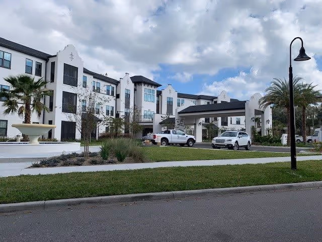 Exterior view of a three-story senior living facility with white walls and dark trim, featuring a covered entrance with two vehicles parked underneath. There are palm trees, a large fountain, a street lamp, and a well-maintained lawn in the foreground under a partly cloudy sky.