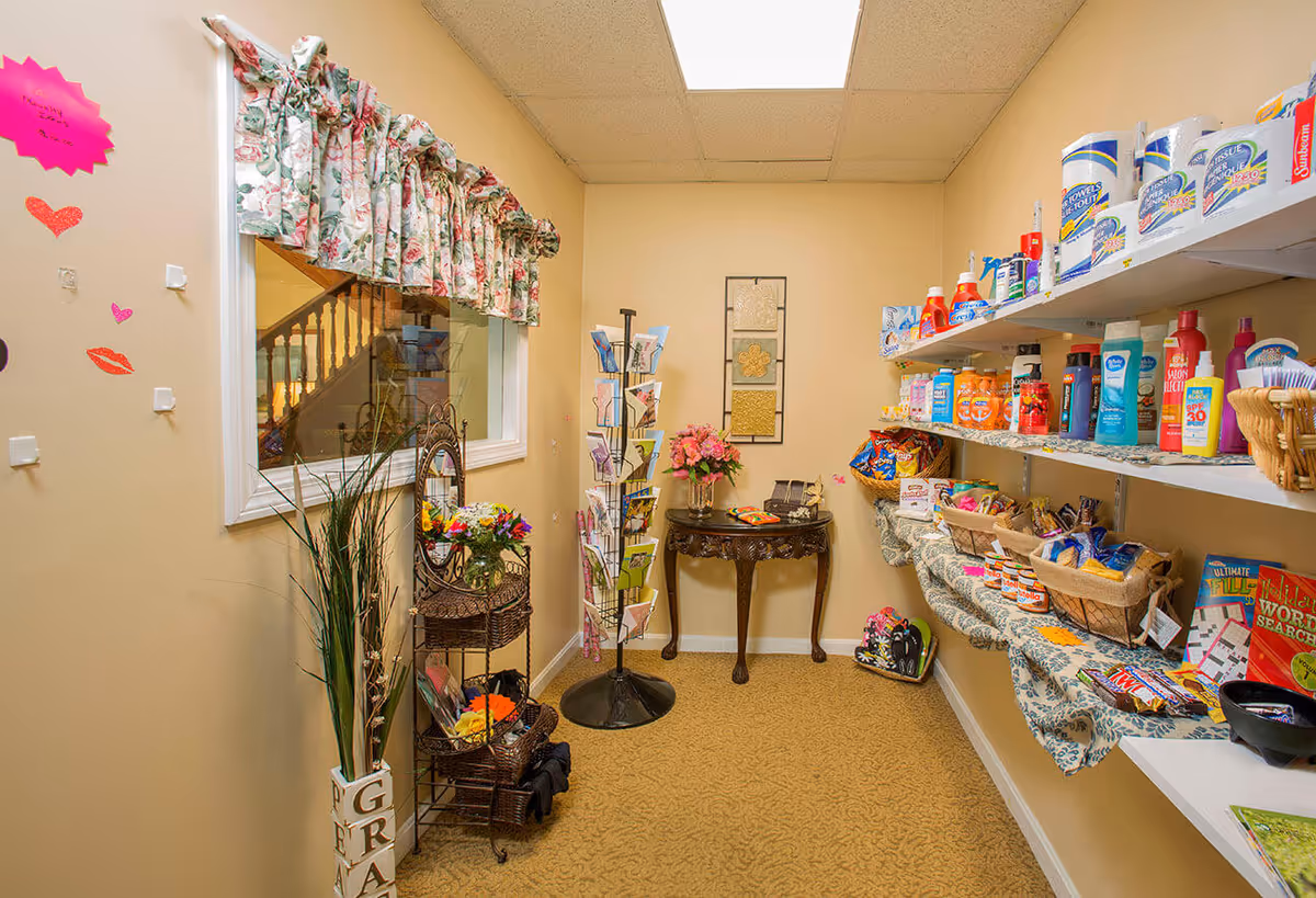 A small retail or convenience area inside a facility with shelves stocked with various personal care products, snacks, and household items. There is a decorative table with flowers at the back, a rotating rack with greeting cards, and a window with floral curtains showing a staircase outside the room. The walls are beige and the floor is carpeted.