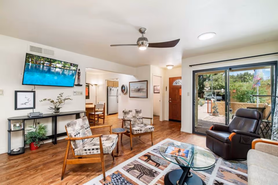 A bright living room with wooden flooring featuring two patterned armchairs, a glass coffee table on a decorative rug, a dark recliner chair, and a beige sofa. A flat-screen TV is mounted on the wall above a console table with plants and framed pictures. Sliding glass doors open to an outdoor patio area with greenery. The room has a ceiling fan and a view into a kitchen and entryway with a wooden door.