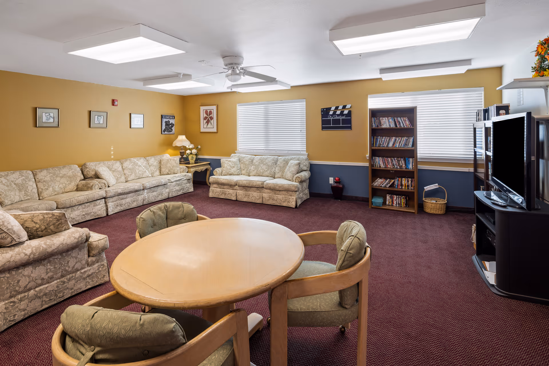 A cozy senior living community lounge with beige floral-patterned sofas arranged along the walls, a round wooden table with four cushioned chairs in the center, a bookshelf filled with DVDs, a flat-screen TV on a stand, and framed artwork on mustard yellow walls with white blinds covering the windows.