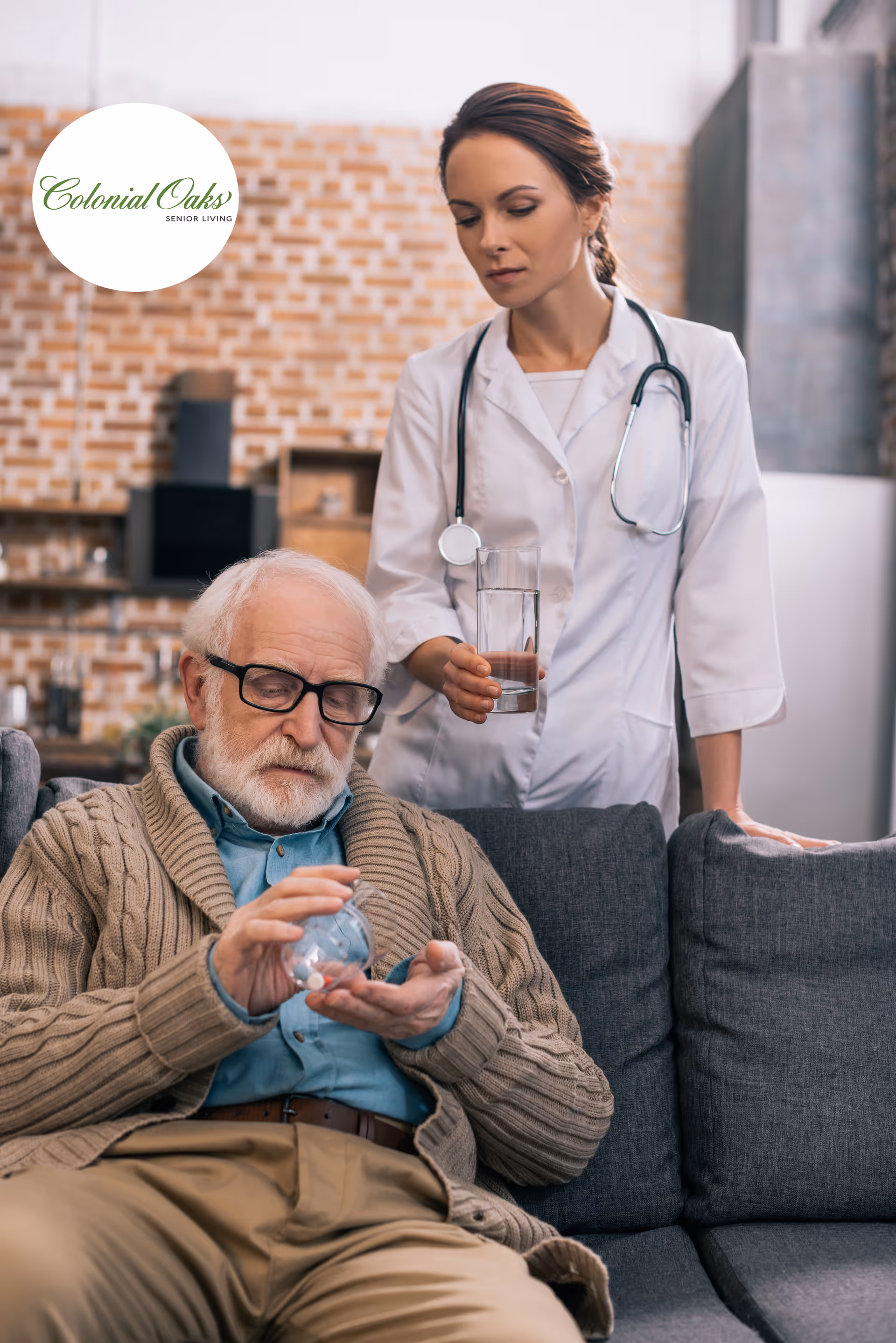 An elderly man sitting on a gray couch, pouring pills from a bottle into his hand, while a female healthcare professional in a white coat with a stethoscope stands beside him holding a glass of water in a cozy living room setting with a brick wall background.