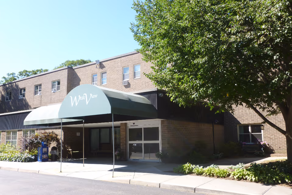 Exterior view of West View Nursing & Rehabilitation Center showing a brick building with a green awning over the entrance. There are trees and shrubs around the entrance area and a clear blue sky above.