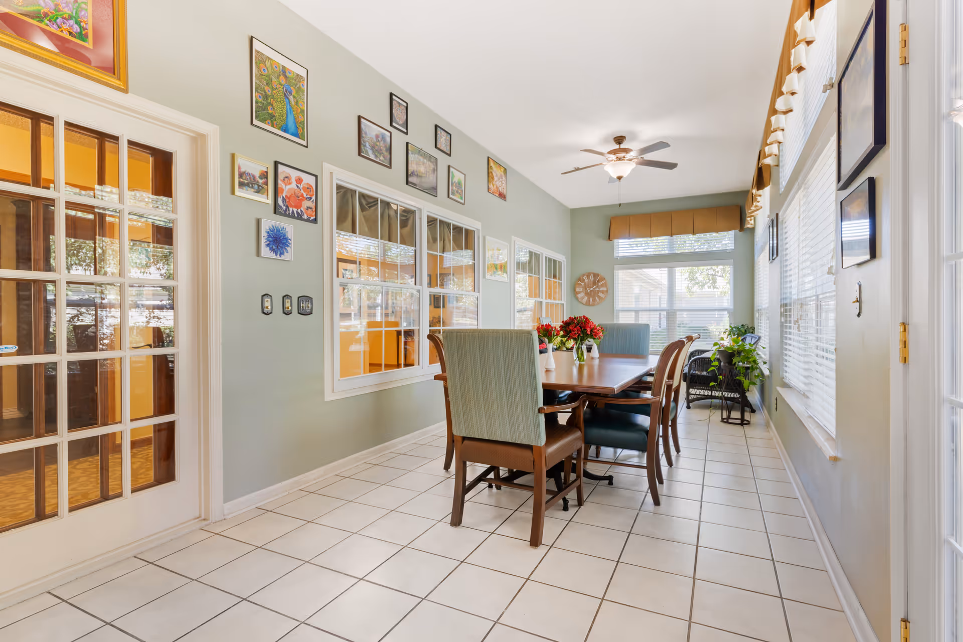Bright dining room with a long wooden table and chairs, tiled floor, large windows, ceiling fan, and wall art.