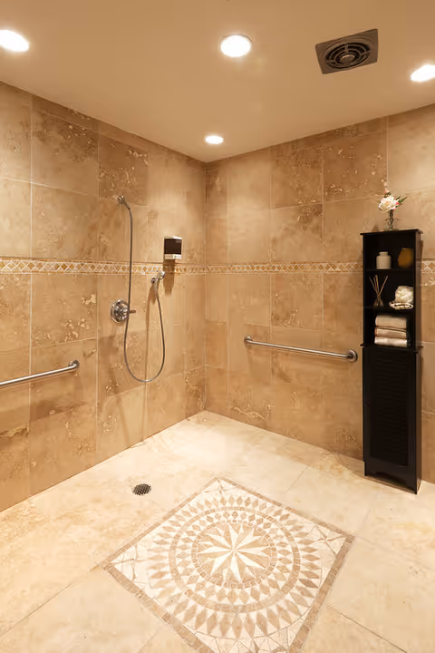 A spacious, tiled shower area with beige marble walls and floor. The floor features a decorative mosaic tile pattern in the center. There are two horizontal grab bars on the walls for support, a handheld showerhead attached to the wall, and a soap dispenser. A black shelving unit stands in the corner holding towels, a flower vase, and other decorative items. The ceiling has recessed lighting and a ventilation vent.