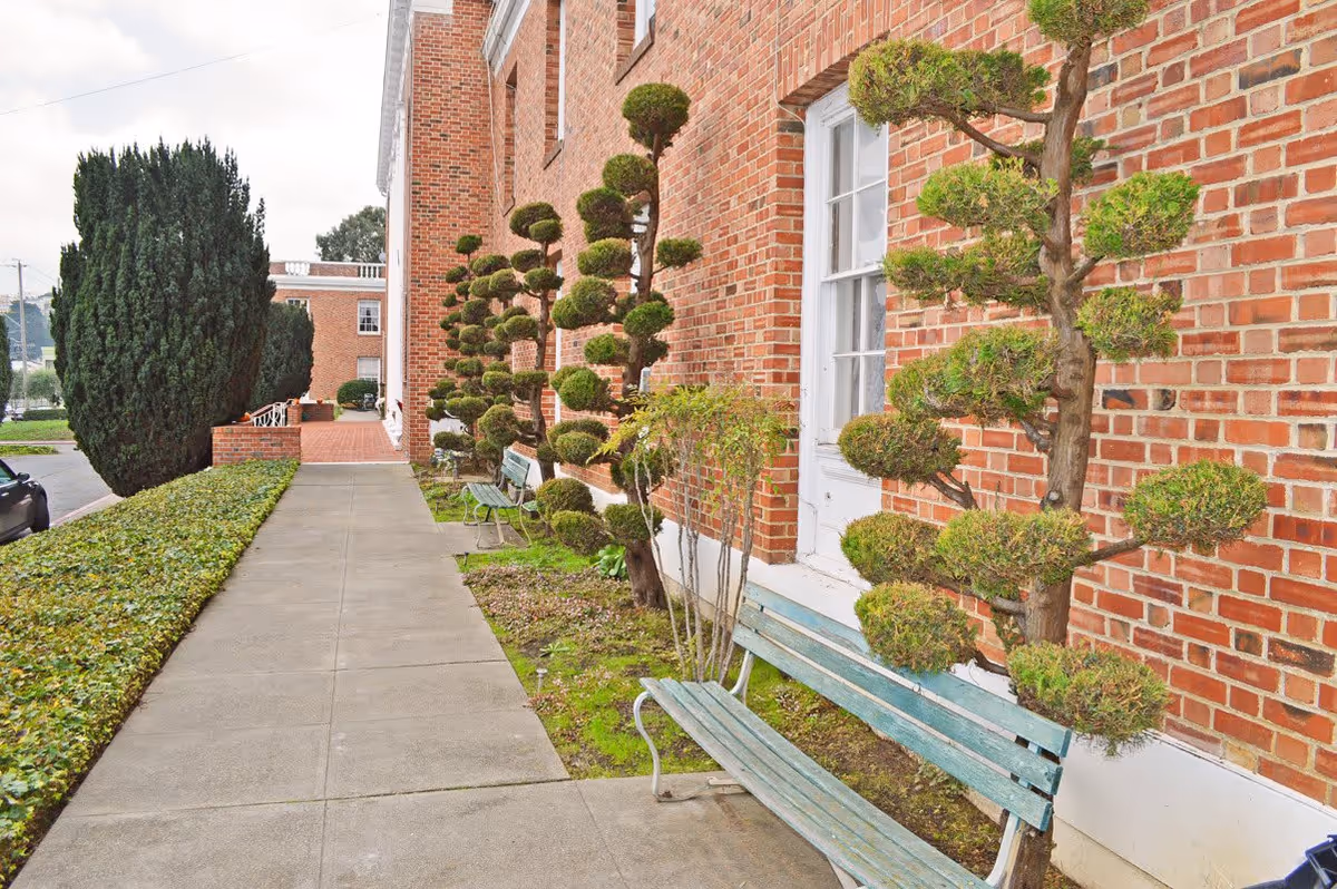 Sidewalk along a red brick building with neatly trimmed topiary trees and green benches placed beside the building. There is a hedge on the left side of the sidewalk and a large bushy tree further down near the street.