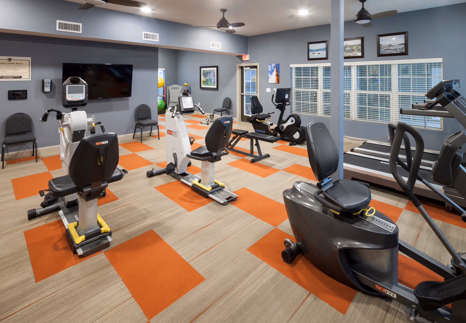 Interior view of a fitness center with exercise equipment including recumbent bikes, treadmills, and weight machines. The room has a patterned carpet with orange and beige squares, gray walls, ceiling fans, framed pictures on the walls, and large windows with blinds.