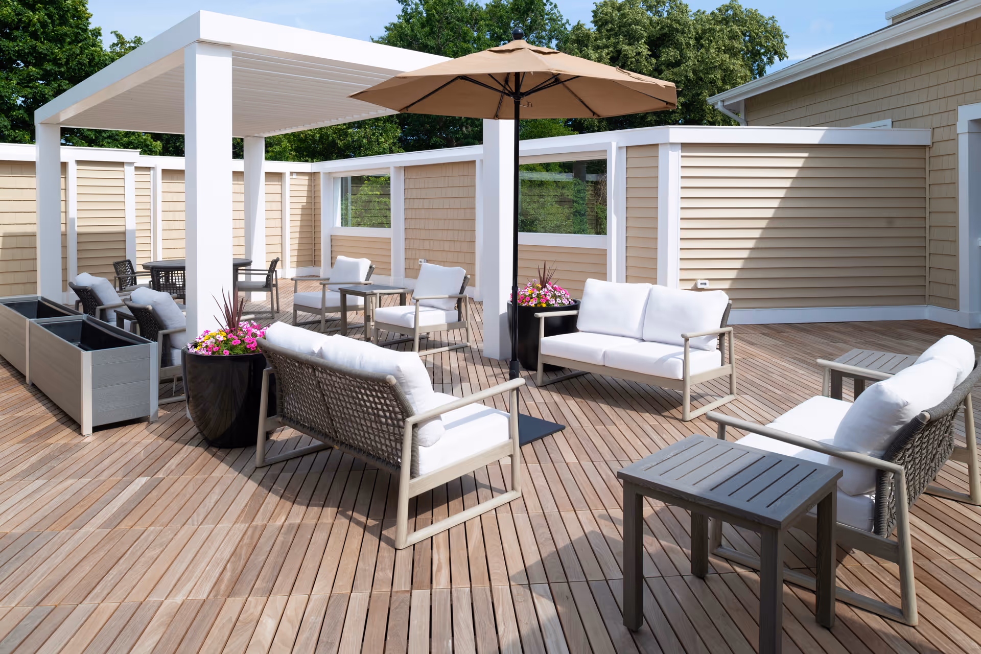 Outdoor patio area with wooden deck flooring, featuring cushioned seating including sofas and chairs arranged around small tables. There is a large beige umbrella providing shade and planters with colorful flowers. The area is enclosed by beige siding walls with white trim and surrounded by greenery.