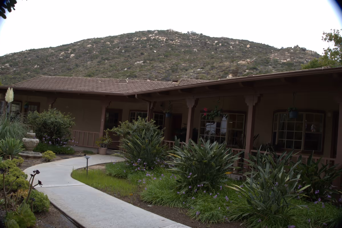 Outdoor view of a senior living facility with a pathway leading through a garden area filled with various plants and shrubs. The building has a covered porch with windows and hanging plants, set against a backdrop of a rocky hillside.