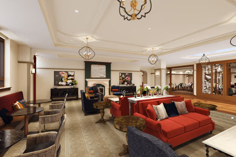A spacious and elegantly decorated living room area with red sofas, multiple armchairs, small round tables, and a fireplace with floral artwork above it. The room features warm lighting from ceiling fixtures and wall sconces, with a carpeted floor and a view into an adjacent dining area through glass doors.