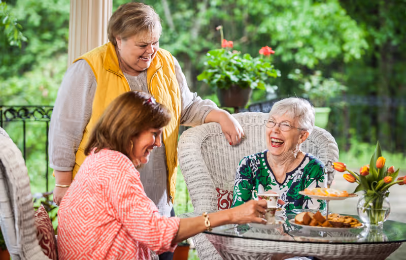 Three women enjoying tea and snacks together on a glass table outdoors. One woman in a yellow vest is standing and smiling, while two other women are seated on white wicker chairs, laughing and holding teacups. There is a vase with orange tulips and a tiered tray with cookies and pastries on the table, with green foliage in the background.
