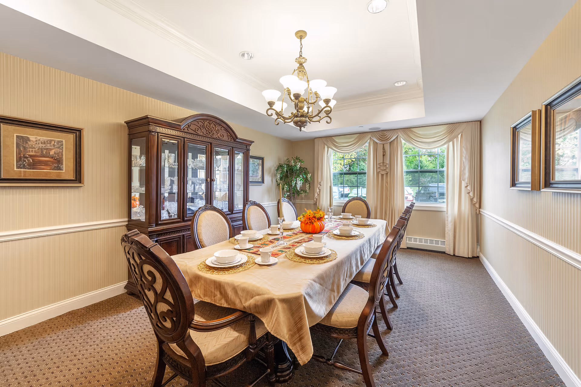A formal dining room with a long rectangular table covered with a beige tablecloth and set with white dishes, cups, and gold placemats. The table has a centerpiece with a small pumpkin and autumn decorations. Surrounding the table are eight ornate wooden chairs with upholstered seats and backs. Against the wall is a large wooden china cabinet displaying glassware. The room has beige walls with white trim, carpeted floor, two large windows with cream-colored curtains, and a brass chandelier hanging from the ceiling.