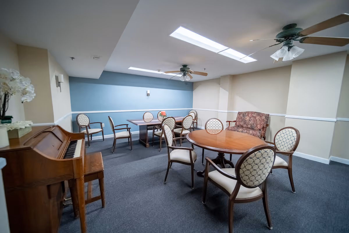 A common room with multiple wooden tables and upholstered chairs arranged around them. There is a wooden piano with a bench on the left side, a floral-patterned loveseat against the wall, and ceiling fans with lights. The walls are painted beige and blue with white trim, and the floor is carpeted in gray.