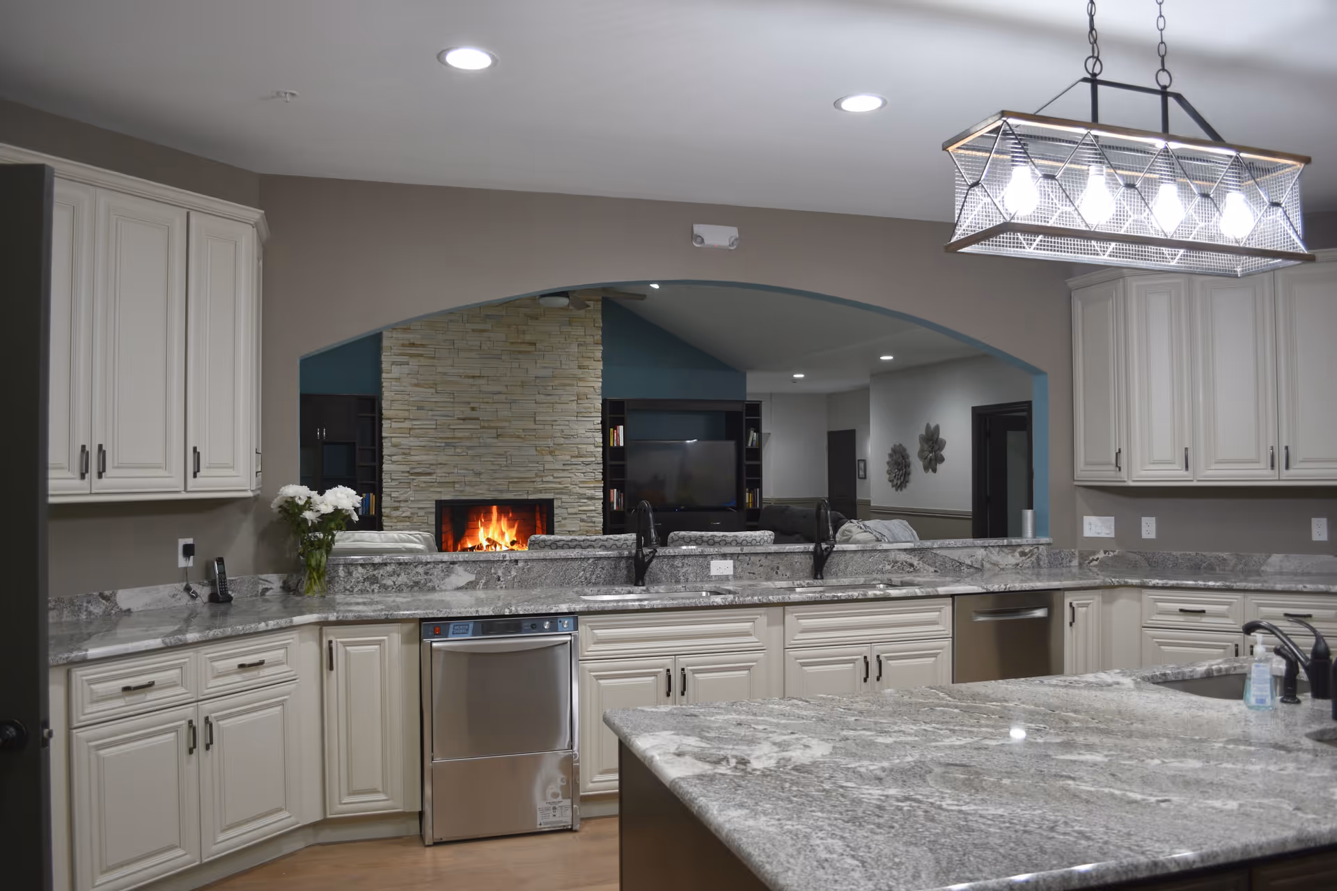 Modern kitchen with white cabinets, granite countertops, a stainless steel dishwasher, and a large island. Through an arched opening, a living room with a stone fireplace and a TV is visible.