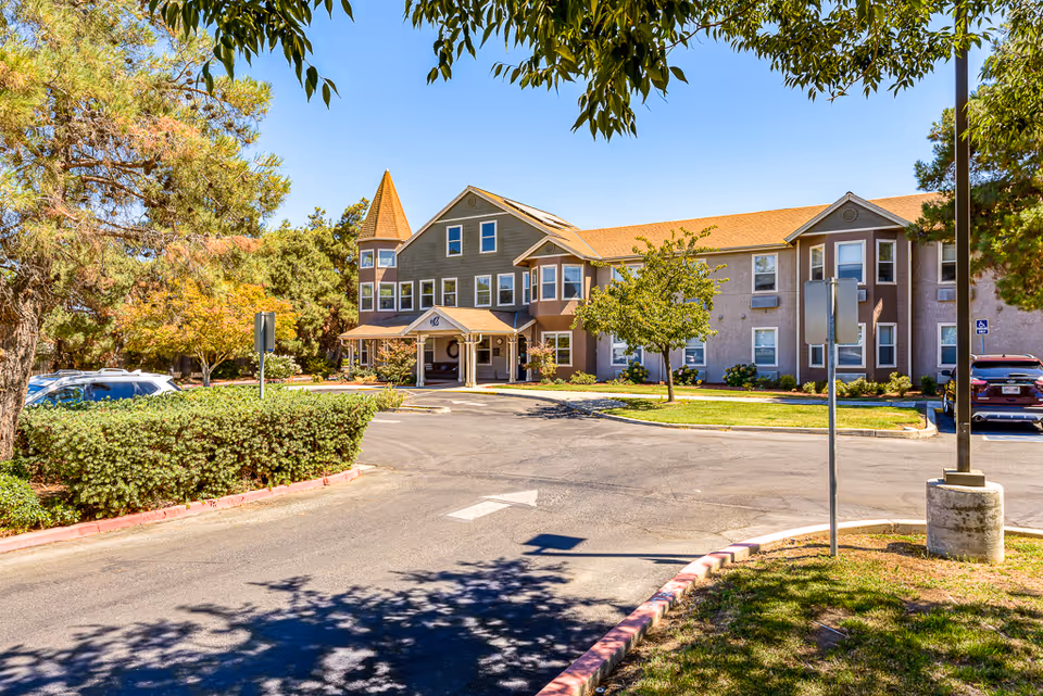 Exterior view of a senior living facility building with a peaked roof and multiple windows, surrounded by trees and greenery under a clear blue sky. There is a driveway and parking area in front of the building with some parked cars.