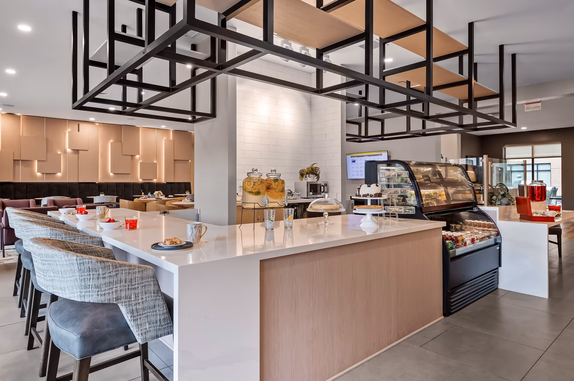 Modern dining area with a large white countertop island featuring several chairs. On the counter are glass beverage dispensers, a cake stand with a cake, cups, and plates with pastries. Behind the counter is a refrigerated display case with various food items. The background shows additional seating with tables and chairs, and a decorative wall with geometric patterns and soft lighting.