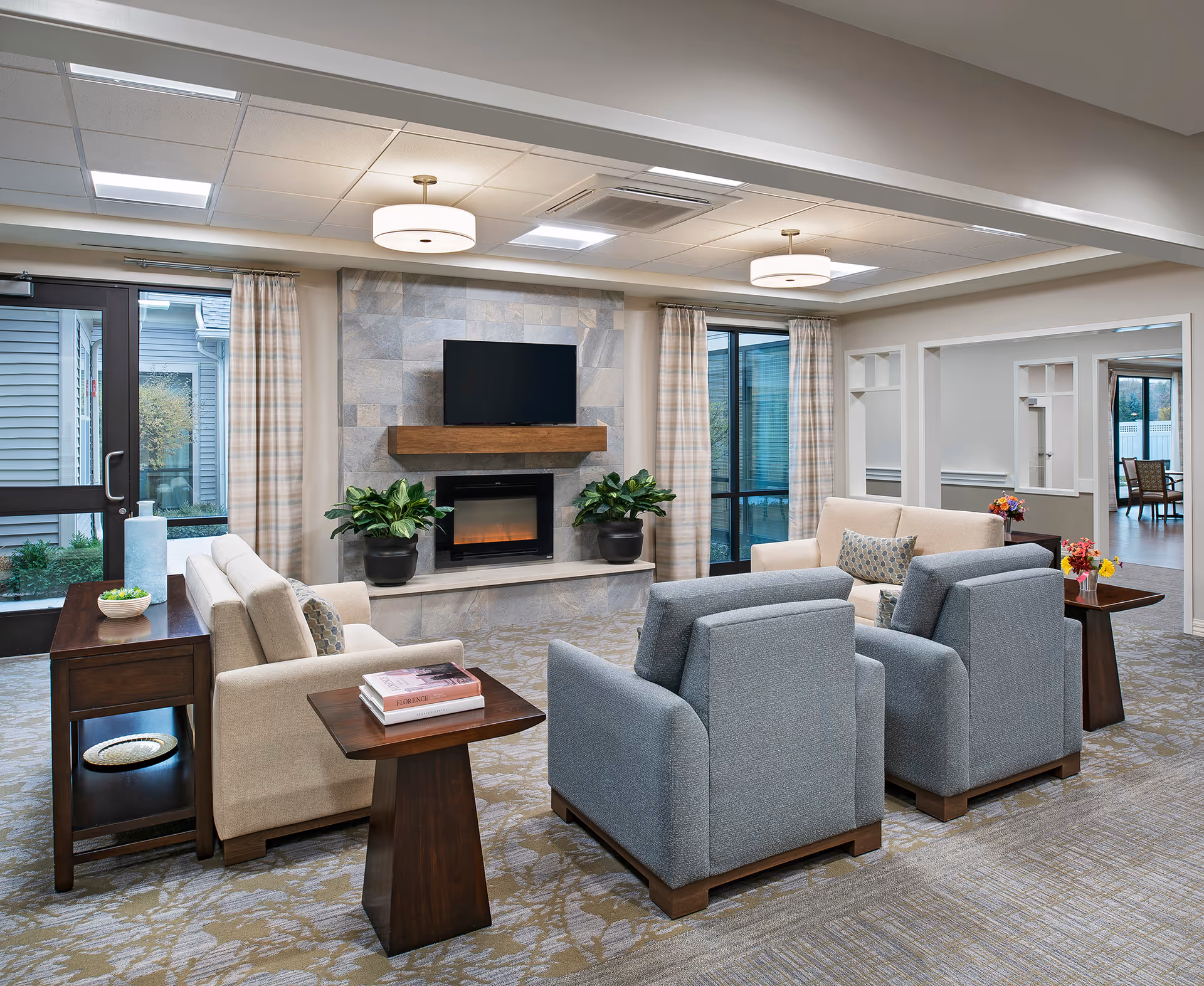 A cozy living room area with two beige sofas and two gray armchairs arranged around a modern electric fireplace with a mounted flat-screen TV above it. The room has large windows with plaid curtains, two ceiling lights, and side tables with books and plants. The carpet has a floral pattern and the walls are painted in neutral tones.