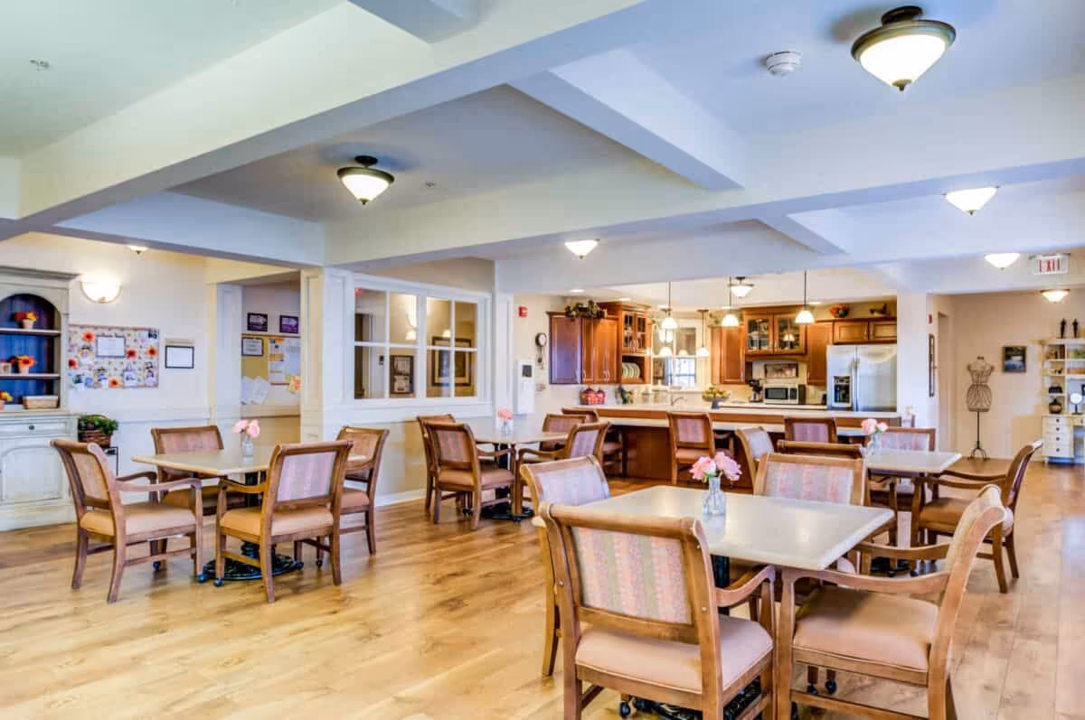 A bright and spacious dining area in a senior living facility with several square tables surrounded by wooden chairs with cushioned seats. The room features light wood flooring, soft lighting from ceiling fixtures, and a kitchen area with wooden cabinets and stainless steel appliances in the background. Small vases with pink flowers are placed on the tables.
