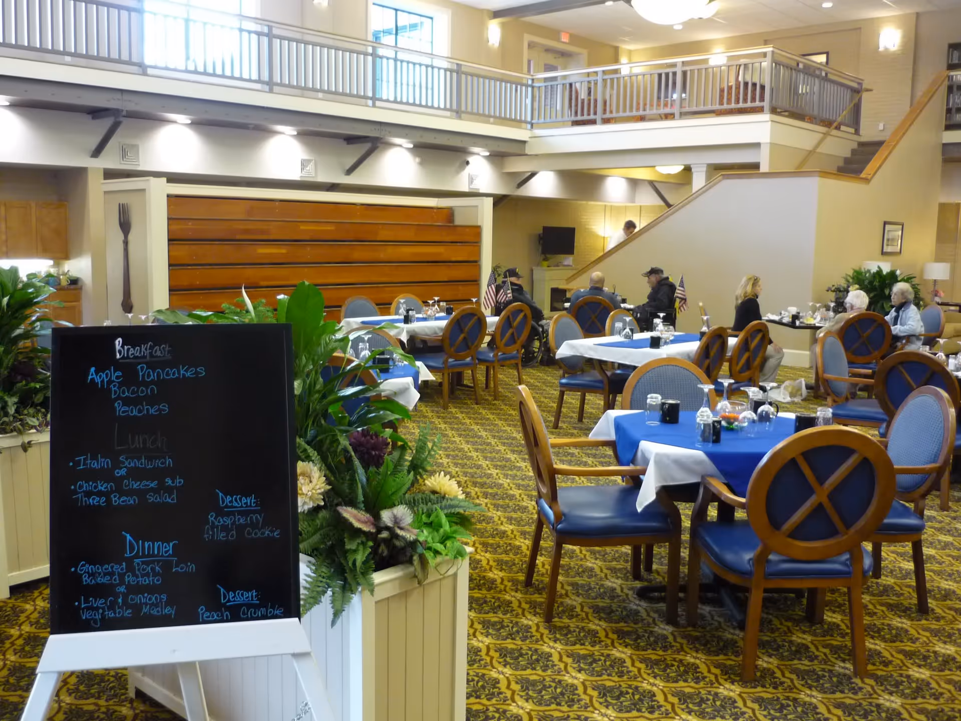 Dining area in a senior living facility with several tables covered in blue and white tablecloths, set with glasses and utensils. A blackboard menu lists breakfast, lunch, and dinner options. Several elderly people are seated and conversing. The room has a patterned carpet, plants, and a second-floor balcony overlooking the area.
