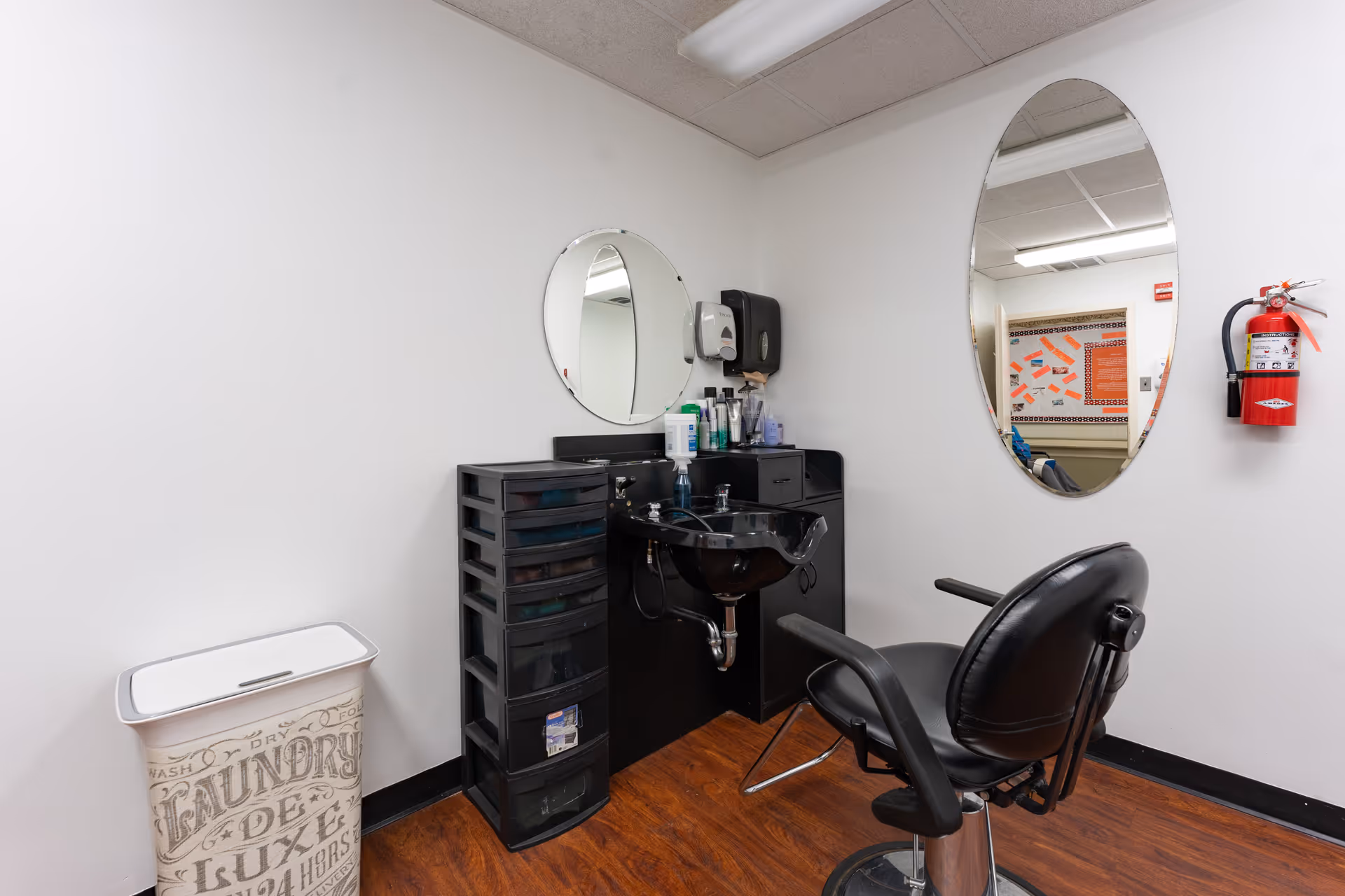 A small salon or grooming area with a black salon chair in front of a black sink and counter. There are two mirrors on the white walls, a black storage drawer unit, various hair care products on the counter, a fire extinguisher mounted on the wall, and a laundry hamper with decorative text on it. The floor is wood-patterned.