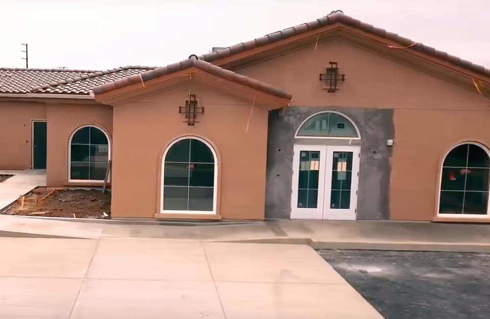 Exterior view of a building under construction with a brown stucco finish, arched windows, and a tiled roof. The entrance features double white doors with an arched window above. The surrounding area includes a concrete walkway and some unfinished landscaping.